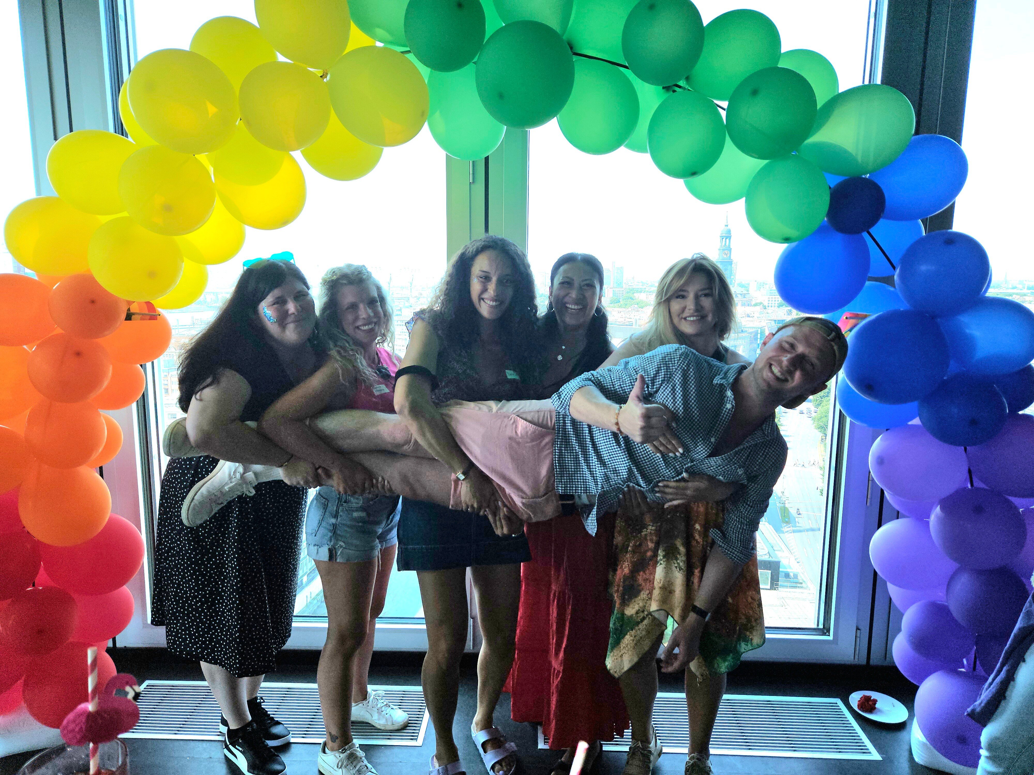 Six people posing under a colorful balloon arch, with one being lifted by the others.