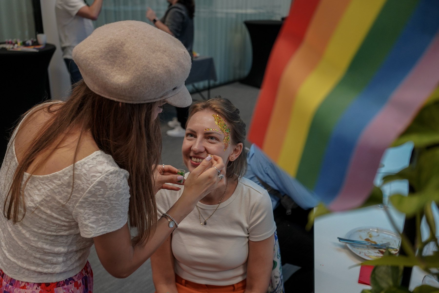 Zwei Personen, eine malt der anderen etwas ins Gesicht. Im Hintergrund ist eine Regenbogenfahne sichtbar.