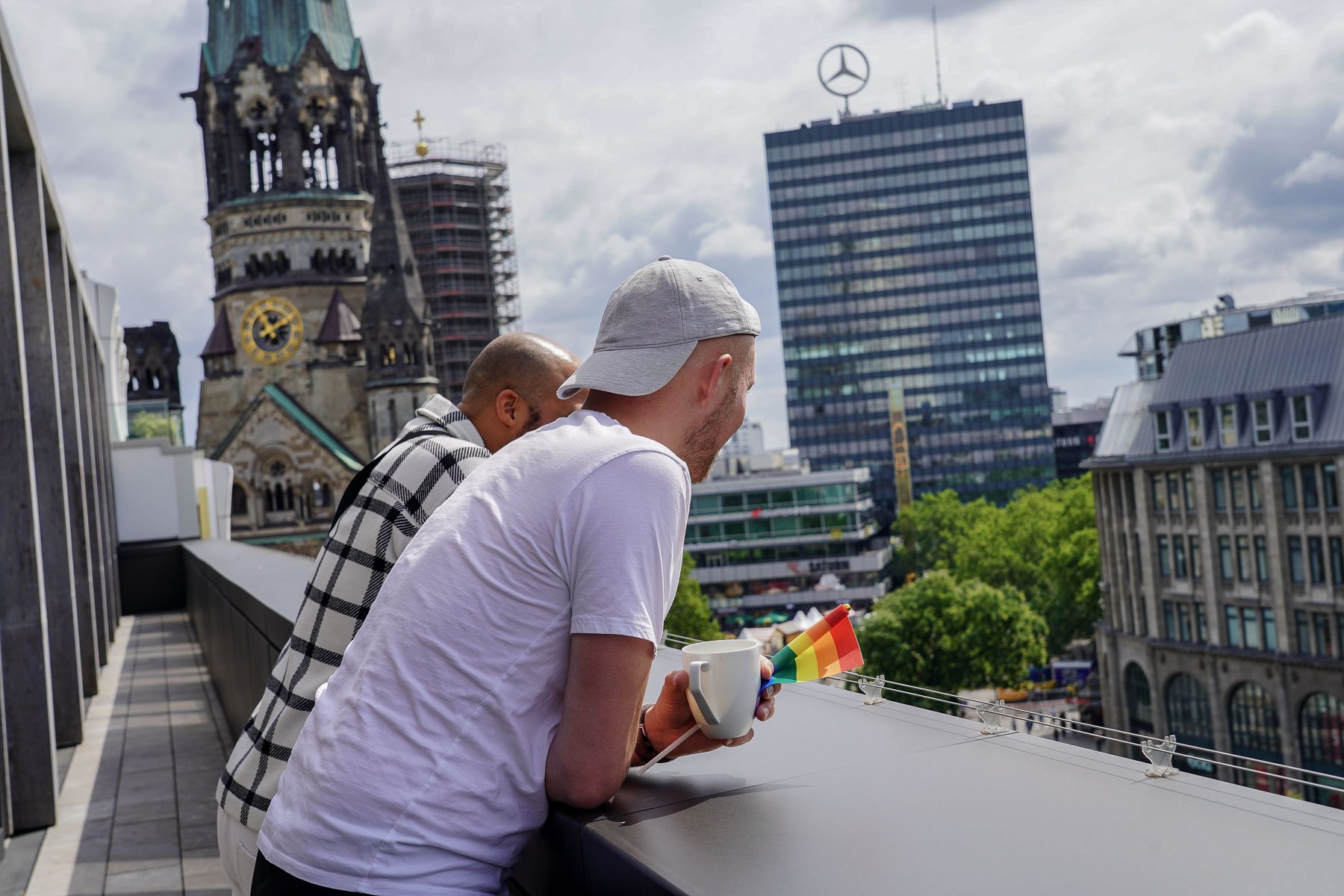  Eine Person steht auf einem Balkon und blickt über die Stadtlandschaft mit einer Regenbogen Flagge.