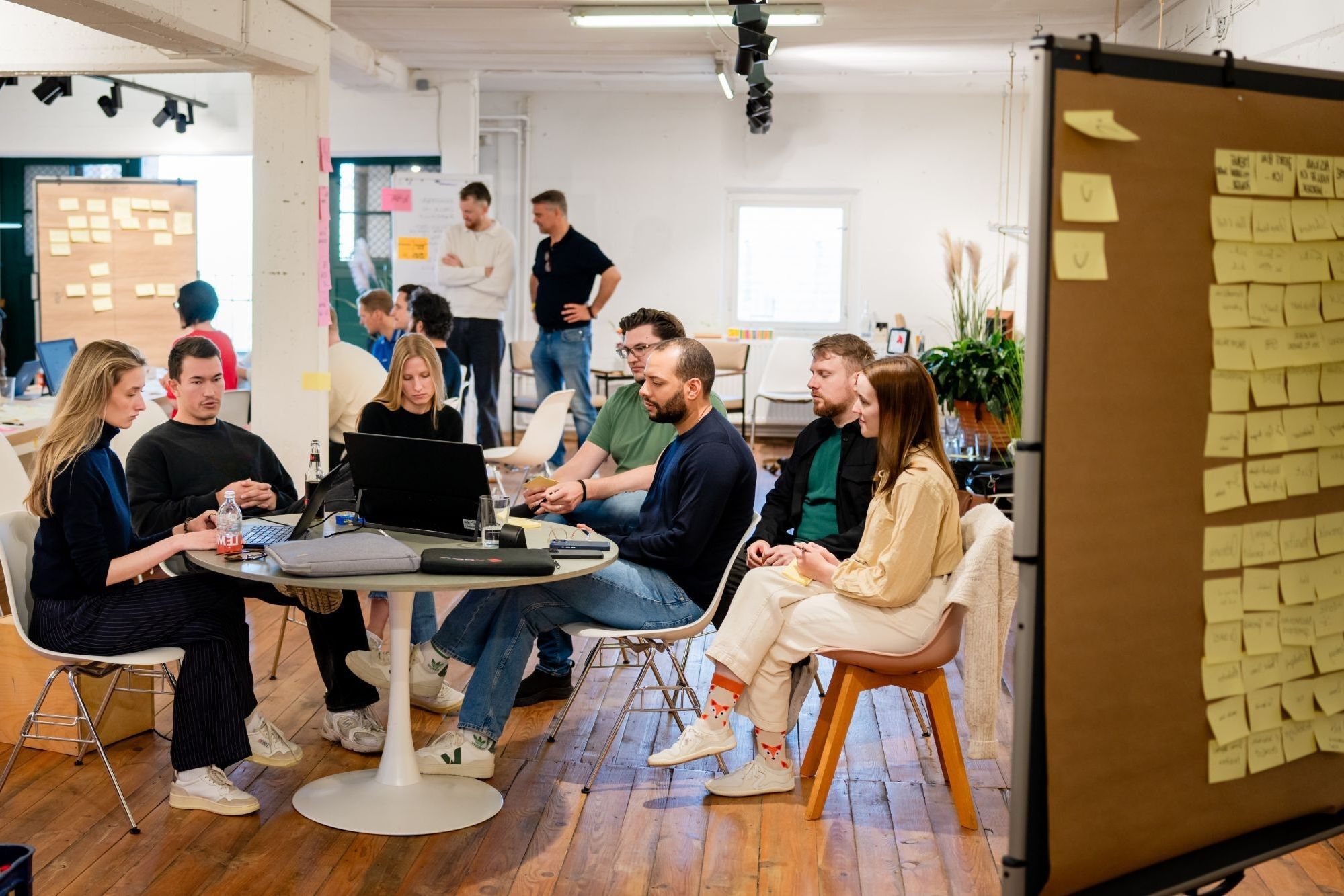 Mehrere Personen arbeiten in einem Workshopraum an Laptops, daneben eine Pinnwand mit Haftnotizen.