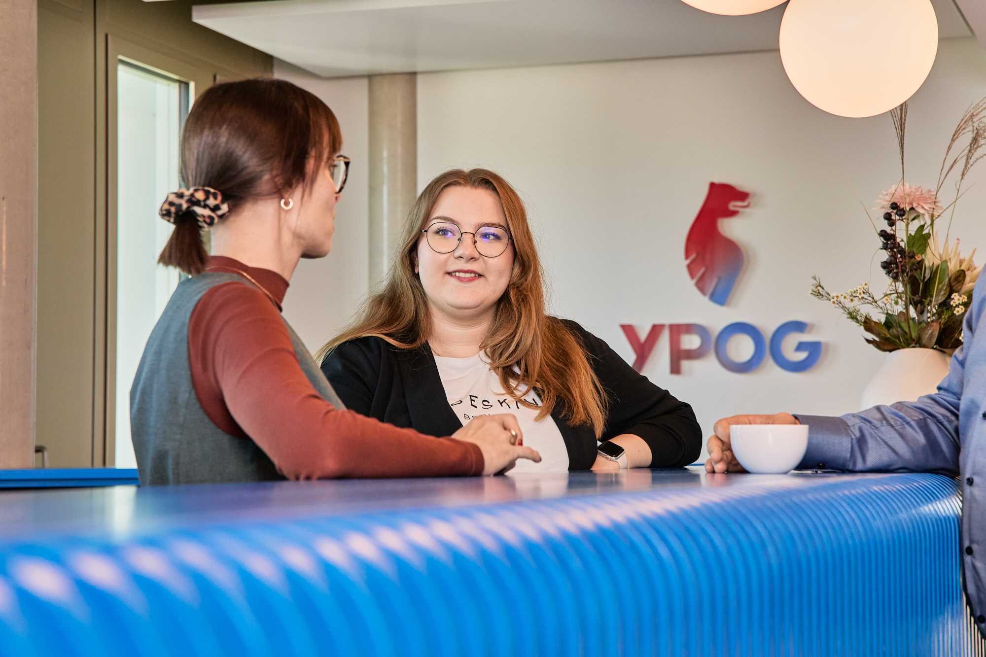 Two people standing at a blue counter in an office having a conversation, with the YPOG logo visible on the wall in the background.